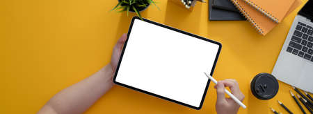 Overhead shot of freelancer writing on blank screen tablet with stylus on yellow worktable with supplies and decorationの写真素材