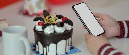 Online Birthday celebration concept, female using blank screen smartphone on white table with cake,...