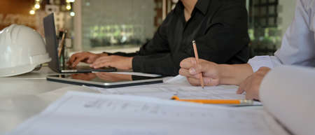 Cropped shot of two engineers working together with blueprint, digital tablet and laptop on office...