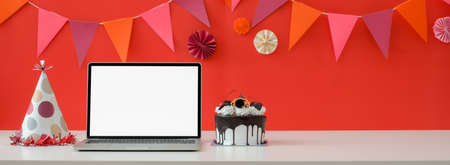 Online Birthday celebration concept with blank screen laptop, cake and copy space on white table with decorations on red wallの写真素材