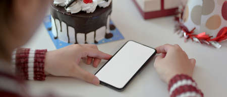 Online Birthday celebration concept, female using blank screen smartphone on white table with cake,...