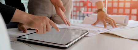 Side view of two engineers consulting on their project with mock-up tablet and paperwork on white table in meeting room の写真素材