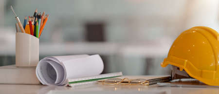 Close up view of engineer worktable with safety helmet, blueprint, measuring tools and other supplies on white desk in glass partition office roomの写真素材