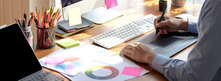 Cropped shot of male graphic designer working on drawing tablet, computer and paperwork on wooden office desk の写真素材