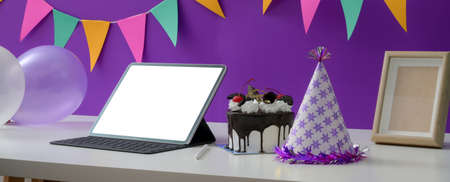Online birthday party concept with blank screen tablet, cake and party hat on white table with decorations on purple wall