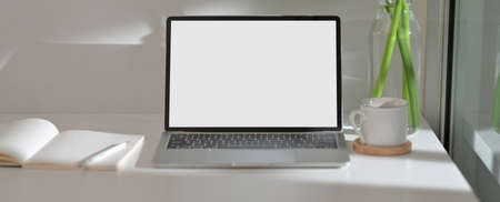 Modern home office desk with mock-up laptop, stationery, cup and plant vase on white tableの写真素材