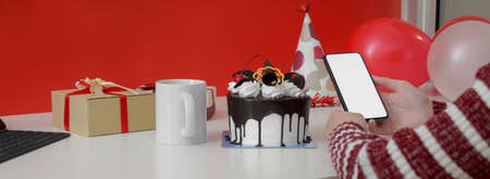 Online Birthday celebration concept, female video calling with smartphone on white table with cake, mug, present boxes and decorationsの写真素材