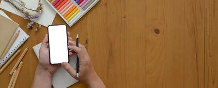 Overhead shot of artist using blank screen smartphone while working on wooden worktable with painting toolsの写真素材