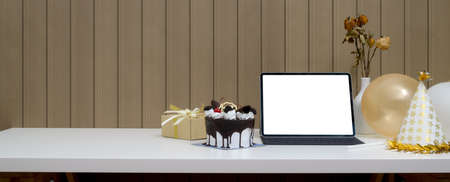 Online birthday party concept with mock-up tablet, Black Forest cake, present box and decorations on white table with plank wallの写真素材