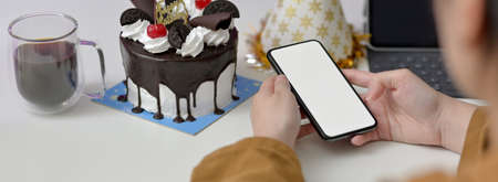 Online birthday party concept, female using mock-up smartphone on white table with cake and party...
