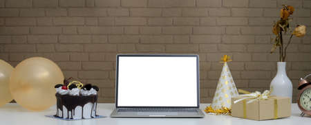 Online birthday party concept with mock-up laptop, Black Forest cake, present box, balloons and party hat on white desk の写真素材