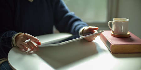 Cropped shot of a woman using digital tablet on white coffee table with book and cup in living room
