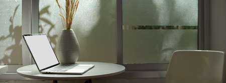 Side view of portable worktable with blank screen laptop and vase on coffee table with chair beside window in room の写真素材
