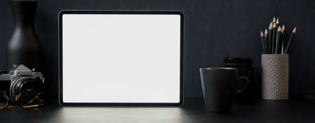Dark modern office desk with blank screen tablet, mug, camera, decoration and other supplies on black tableの写真素材