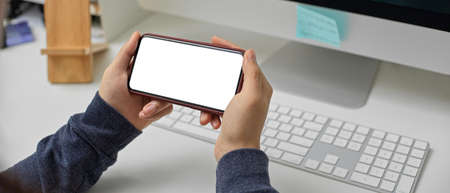 Cropped shot of female employee relaxing with horizontal mock-up smartphone while working with computer on office desk の写真素材