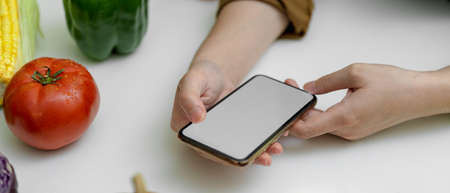 Cropped shot of female looking recipe on mock-up smartphone while preparing ingredient for cooking on kitchen table with fresh vegetablesの写真素材