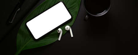 Top view of blank screen smartphone, wireless earphone above leaf and coffee cup on dark backgroundの写真素材