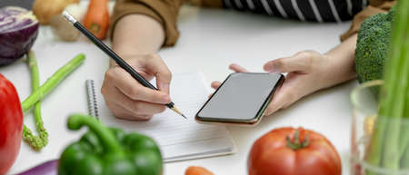 Cropped shot of female noting ingredient on notebook while looking information on mock-up smartphone...