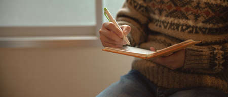 Close up view of a girl writing on diary notebook while sitting at reading corner beside window