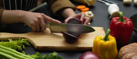 Side view of female cook cutting fresh red onion on chopping block at dark modern kitchen 