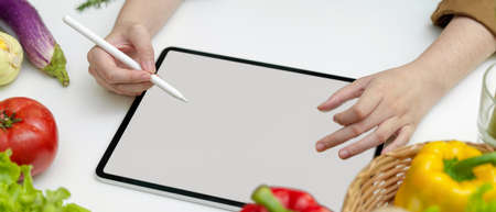 Cropped shot of female searching menu for dinner on mock-up tablet on white kitchen table with fresh...