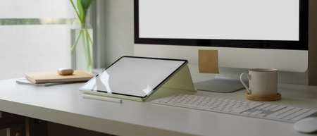 Cropped shot of simple office desk beside window with mock-up computer, digital tablet, schedule books and decorationの写真素材