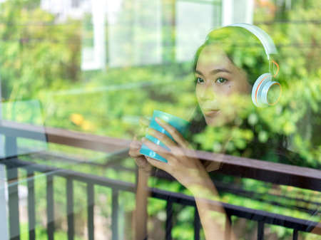 Through glass window view of relaxed woman listening to music with headphones and holding a coffee...