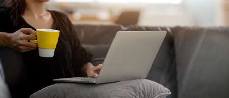 Cropped shot of a woman relaxing with computer laptop and holding hot coffee cup while sitting on couch in living room の写真素材