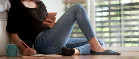 Close up view of female freelancer taking note on schedule book and smartphone while sitting on floor at bedroomの写真素材