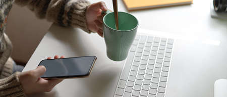 Close up view of female employee relaxing with smartphone and holding a cup of coffee at office desk...