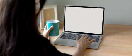 Cropped shot of female searching informations on mock-up laptop and holding mug at home office desk の写真素材