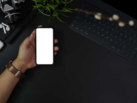 Overhead shot of hand holding blank screen smartphone above dark modern office desk with office supplies and decorationsの写真素材