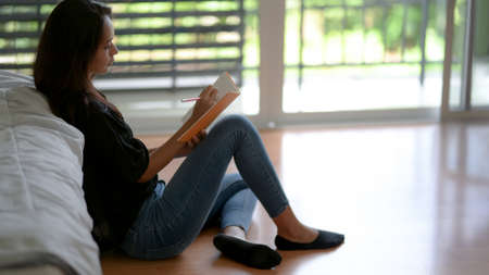 Cropped shot of young female noting on blank notebook while sitting on the floor near bed の写真素材