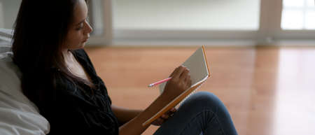 Close up view of young female writing on blank schedule book while sitting on the floor near bed