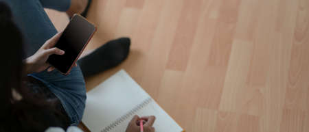 Overhead shot of young female noting on blank notebook and looking information on smartphone while...