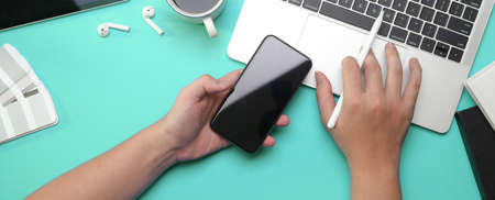 Overhead shot of male freelancer left hand holding smartphone and right hand typing on laptop
