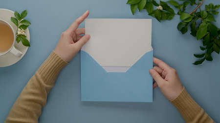 Overhead shot of female hands open mock-up greeting card with blue envelope on table with tea cup and leaves の写真素材