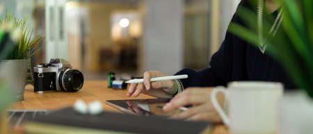 Cropped shot of female photographer working on digital tablet with stylus pen on wooden table with...