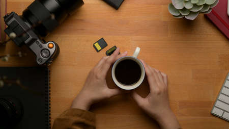 Overhead shot of female hands holding coffee cup on wooden worktable with office suppliesの写真素材