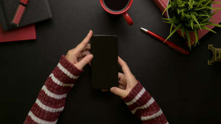 Overhead shot of female using smartphone while sitting at stylish office desk with cosmetics, supplies, coffee cup and decorationの写真素材