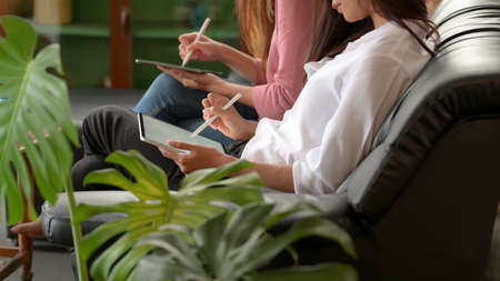 Side view of female college students relaxed sitting on black couch and using digital tablets with...