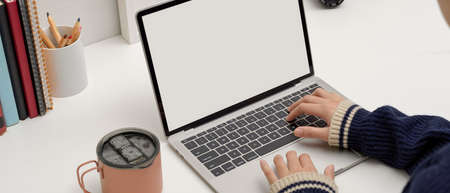 Cropped shot of female freelancer typing on mock-up laptop on white desk with ice coffee mug, books...