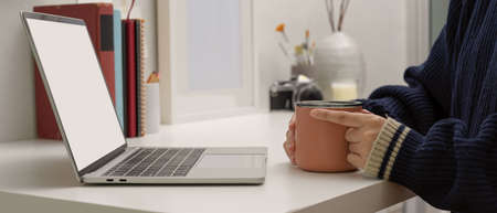 Side view of female freelancer sitting at home office and holding coffee mug while looking on mock...