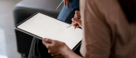 Cropped shot of female using mock up tablet with stylus pen while sitting on sofa in living room