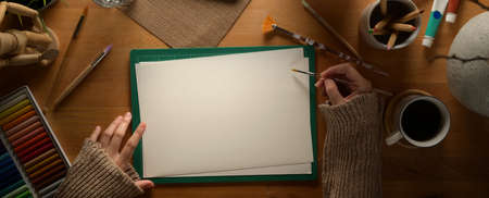 Overhead shot of female artist drawing on sketch paper on wooden worktable with painting tools and...