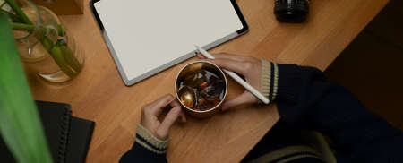 Overhead shot female hands holding ice coffee mug and stylus pen while working with mock up tablet...