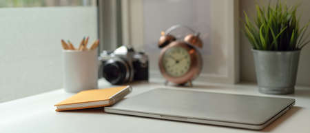 Close up view of study table with closed laptop, notebook, stationery, camera, clock and decorations on white tableの写真素材