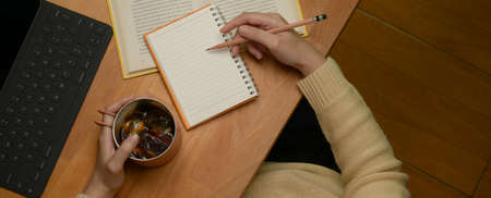 Overhead shot of female writing diary notebook on study table with coffee cup, tablet and book