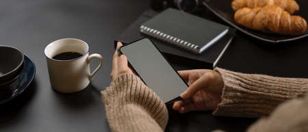 Side view of female take a break with mock up smartphone while sitting at workspace with supplies, coffee cup and croissantの写真素材