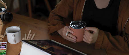 Cropped shot of female artist holding coffee cup while working with tablet on worktable with...